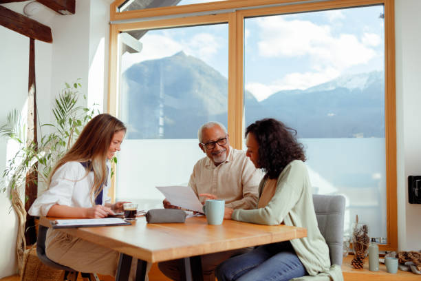 A woman, likely a financial advisor, is seated with a senior couple in a bright room with a stunning mountain view, indicative of a home in Slovenia. They are engaged in conversation, with documents laid out on the table, pointing to a discussion about pension insurance. The advisor is presenting information, while the couple listens intently, considering the options for their retirement planning.